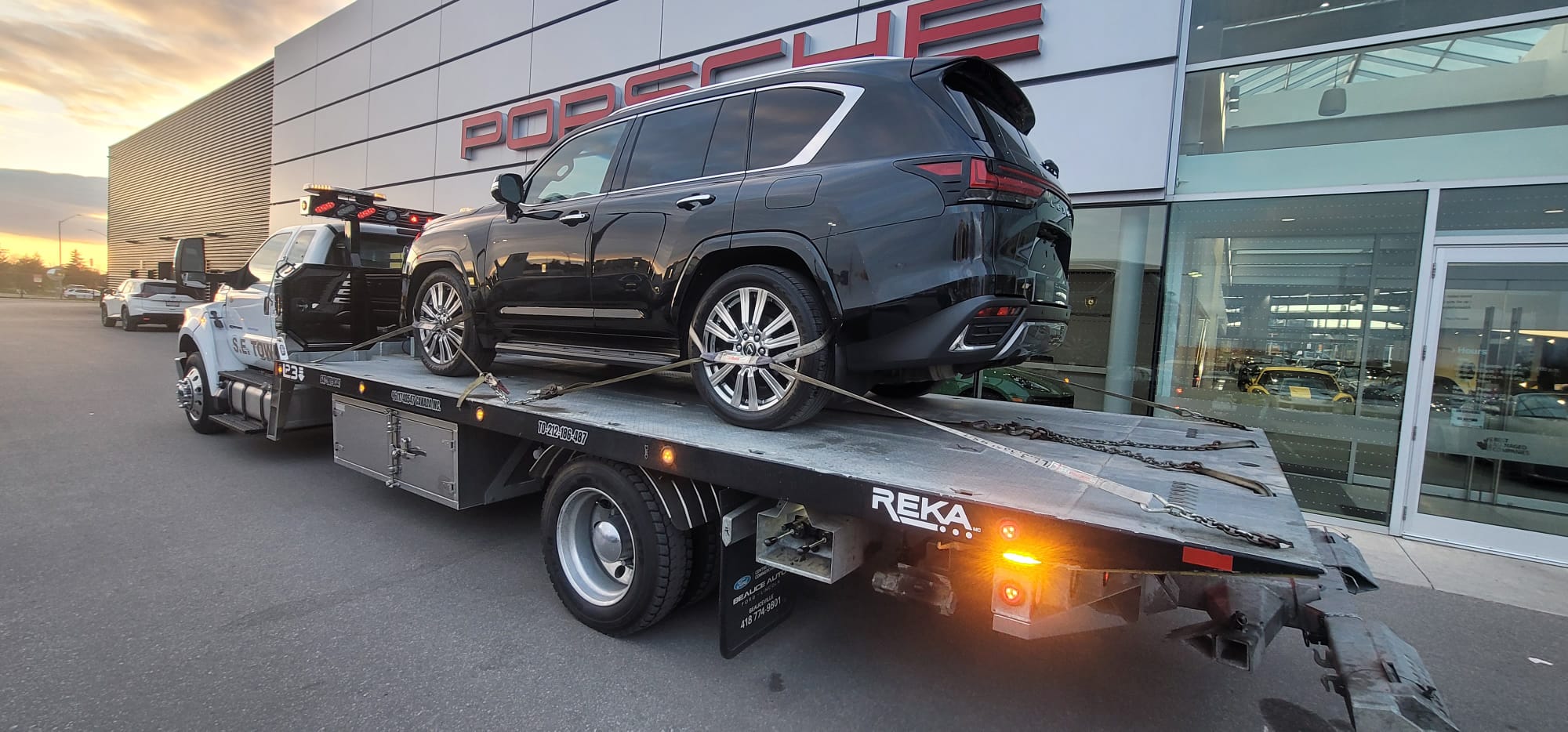 Black Lexus LX 600 luxury SUV loaded on S.E. Towing flatbed at Porsche dealership at sunset